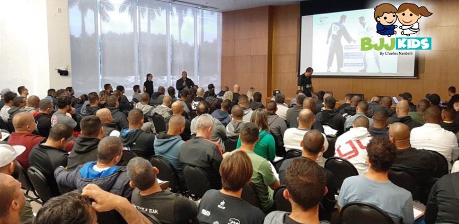 Auditório lotado de professores em formação BJJ Kids, com a marca projetada na tela