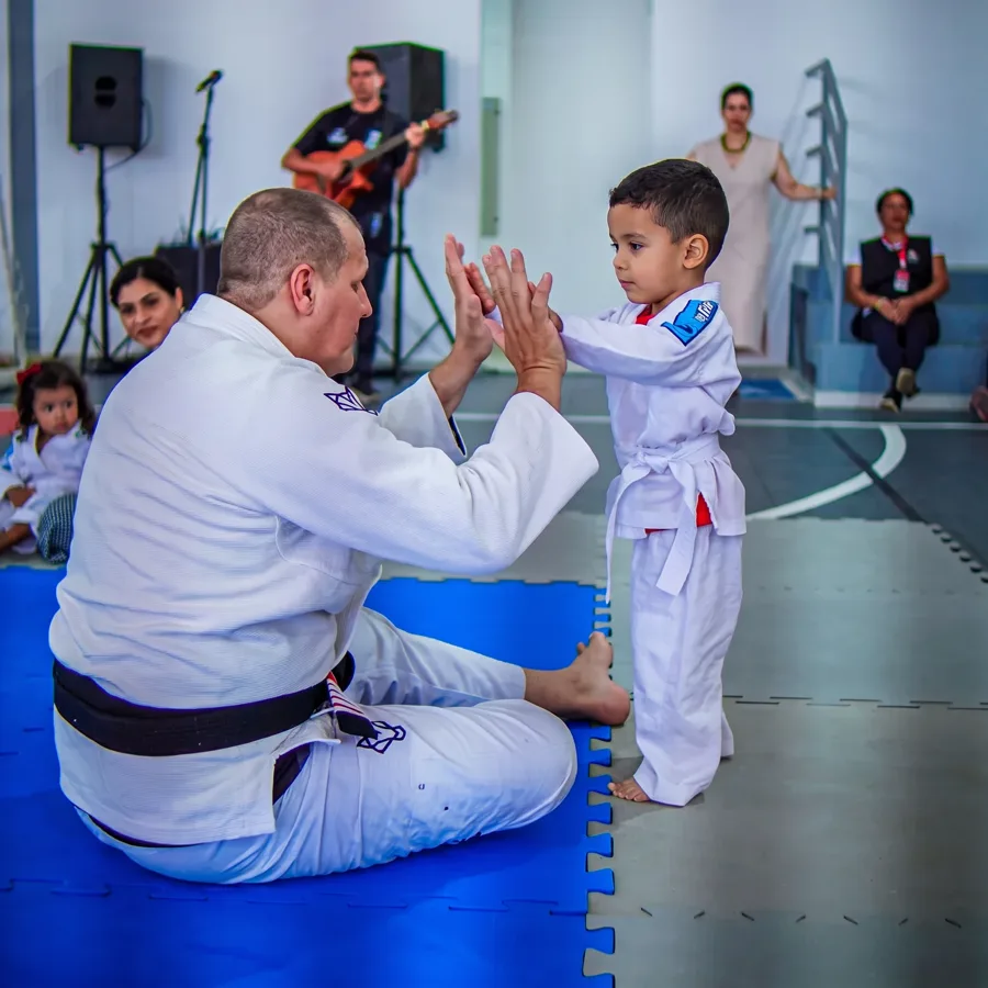 Charles sentado num tatame azul, de kimono e faixa preta, com as mãos encostando nas de um menino pequeno de kimono em pé à frente dele